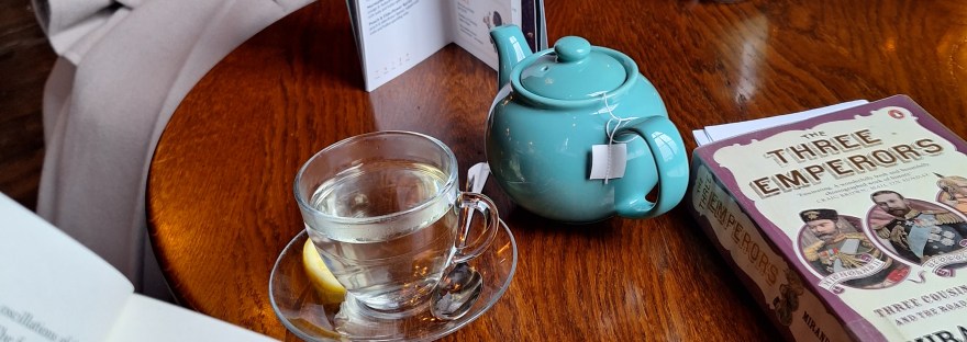 reading a book whilst sitting at a table with a cup of tea, a teapot, and a book on it